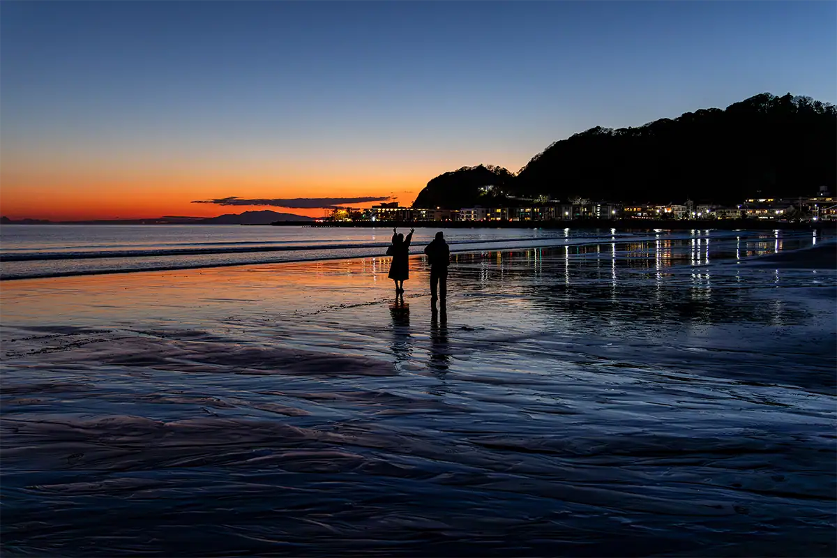 Two people standing on a reflective beach at sunset with coastal hills and city lights in the distance.