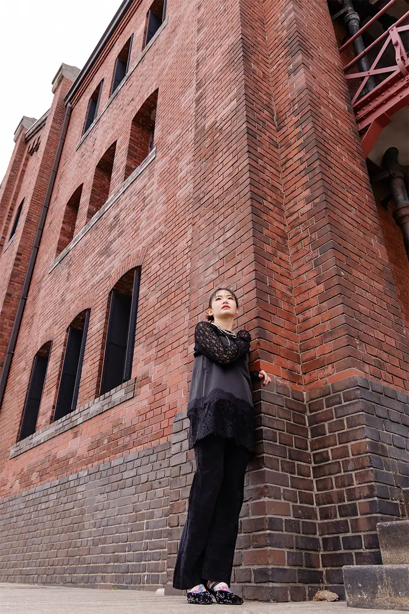Travel portrait of a woman beside a tall red-brick building, photographed with the Tamron 17-70mm lens for Canon RF mount.