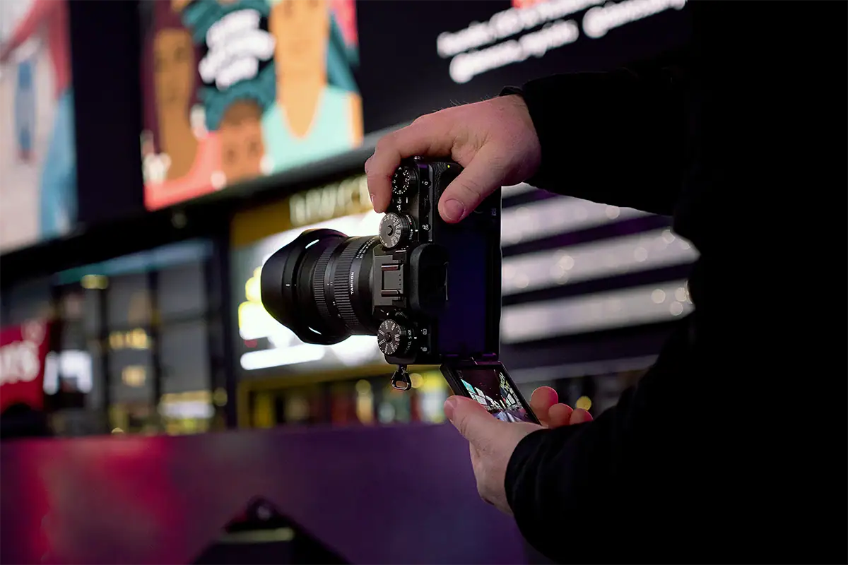 Photographer adjusting a mirrorless camera while shooting handheld in a brightly lit city at night.