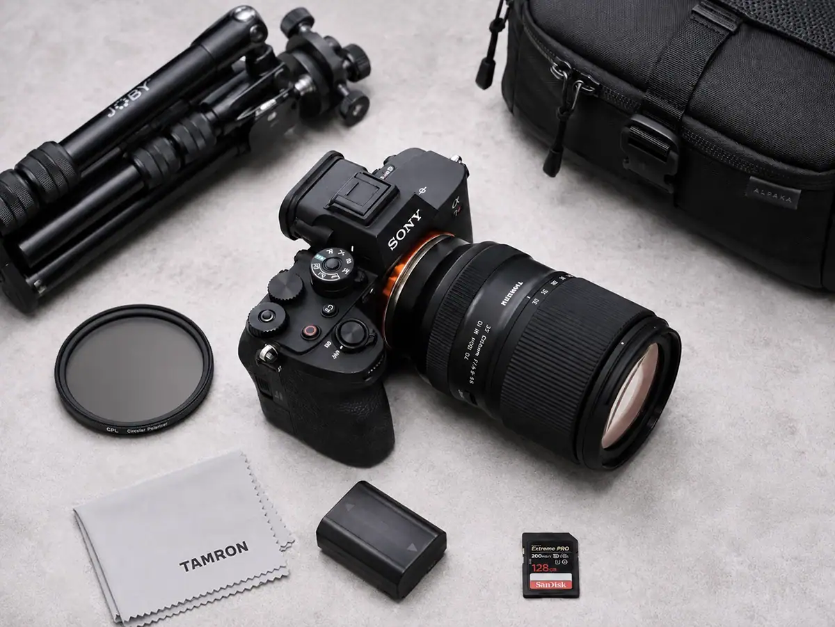 Travel photography gear lay flat with a mirrorless camera, Tamron zoom lens, compact tripod, polarizing filter, battery, memory cards, lens cloth, and camera bag.