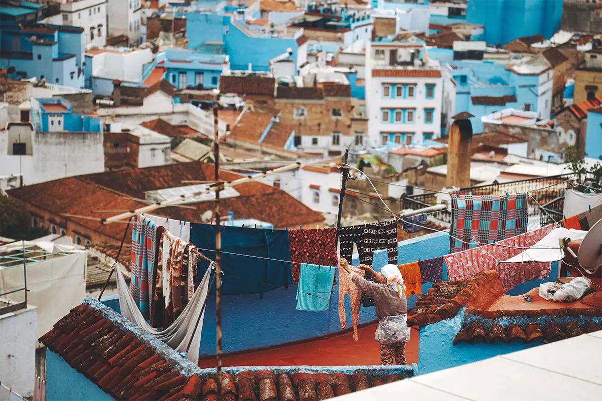 Woman hanging colorful textiles on a rooftop overlooking the blue-painted buildings of Chefchaouen, Morocco.