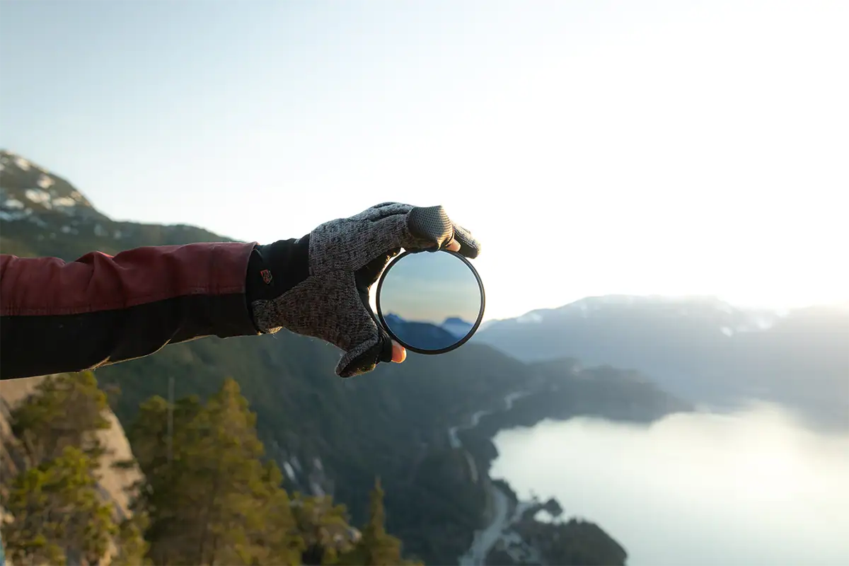 Hand holding a circular polarizer filter against a mountain landscape to demonstrate glare control and improved outdoor image contrast.