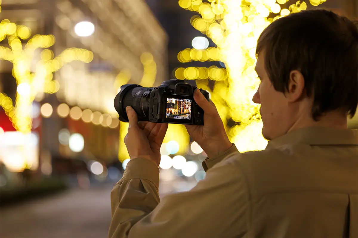 Fotógrafo usando uma câmera sem espelho com uma lente zoom padrão para compor uma cena noturna da cidade com luzes decorativas ao fundo.