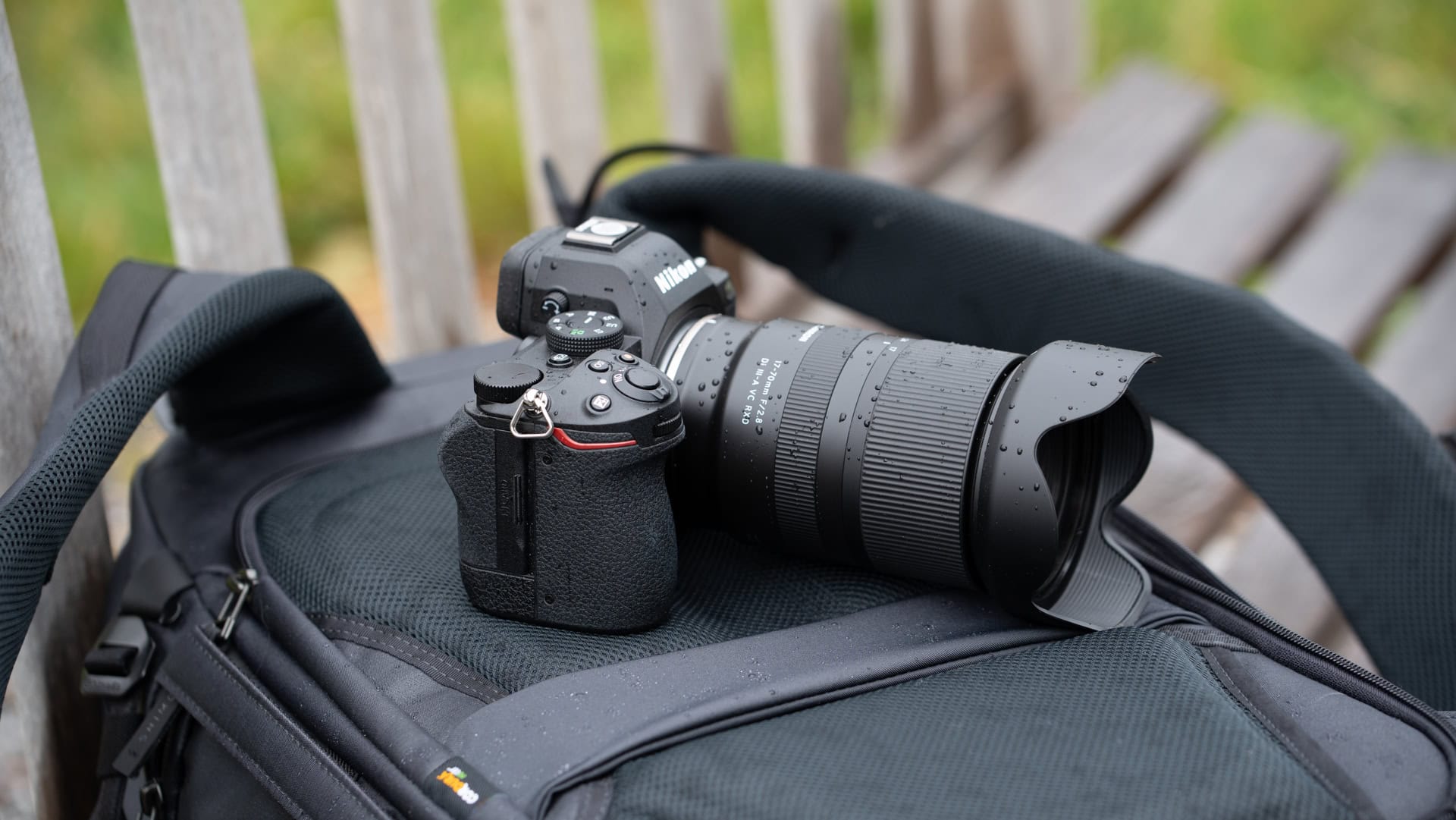 Câmera sem espelho da Nikon com uma lente grande-angular apoiada em uma mochila ao ar livre, com gotículas de água visíveis na câmera e na lente.