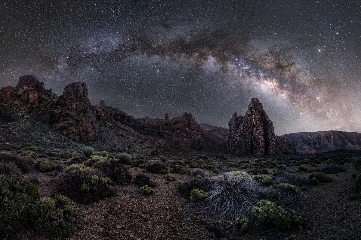 Milky Way stretching across a star-filled night sky above a rugged rocky landscape.