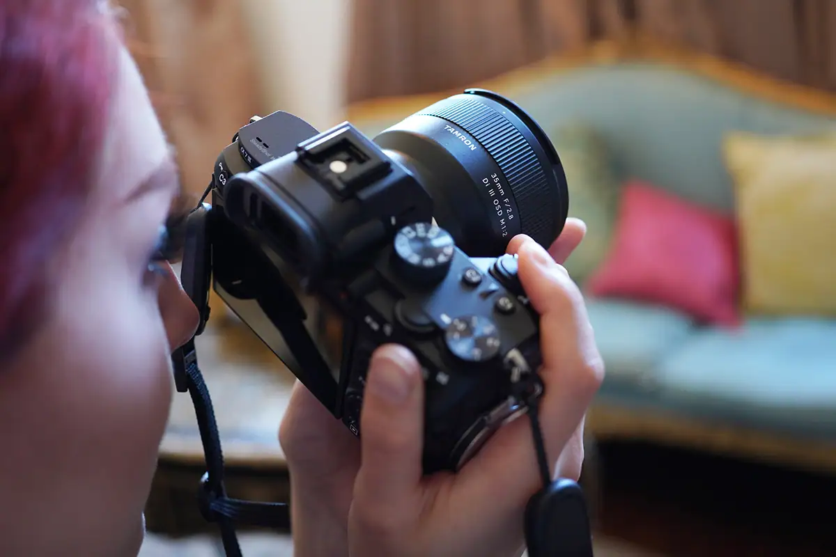 Photographer holding a camera fitted with a compact Tamron 35mm prime lens while reviewing the composition on the rear screen.
