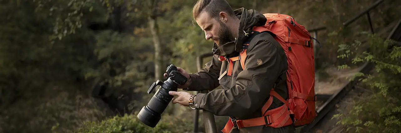 Outdoor photographer carrying a backpack while adjusting a camera fitted with a Tamron telephoto zoom lens.