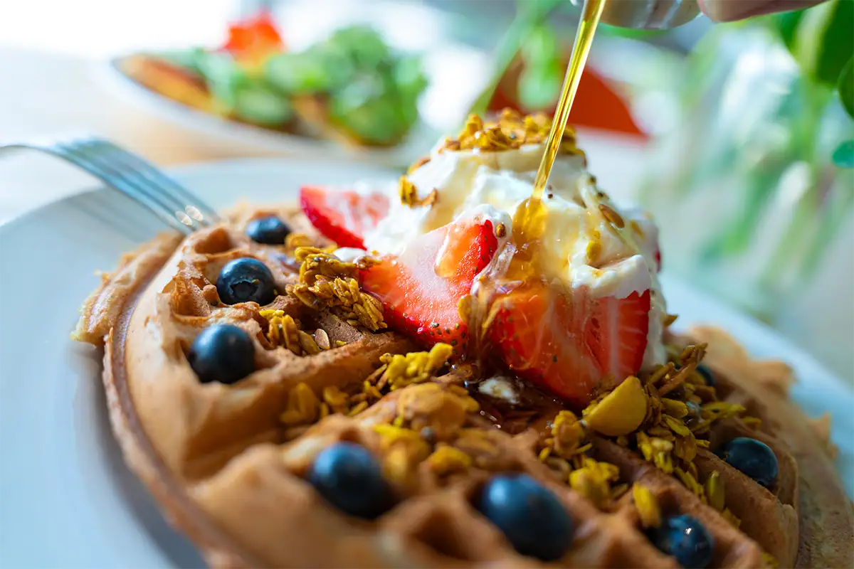 Close-up de um waffle coberto com morangos, mirtilos, iogurte, granola e calda, fotografado com uma lente zoom padrão para destacar os detalhes contra um fundo suavemente desfocado.