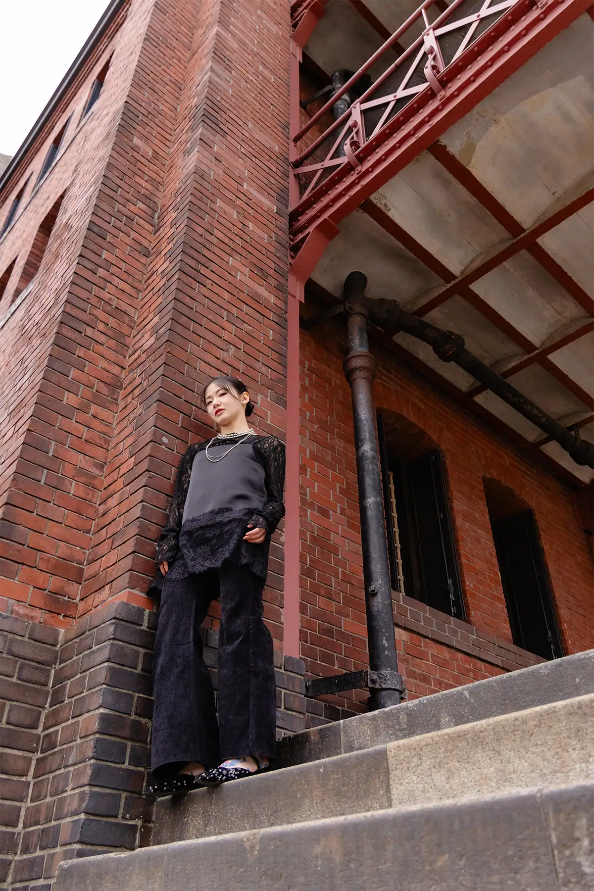 Retrato ambiental de uma mulher em pé nos degraus ao lado de um prédio de tijolos vermelhos em estilo industrial, fotografada com uma lente zoom padrão para incluir tanto a pessoa quanto a arquitetura ao redor.