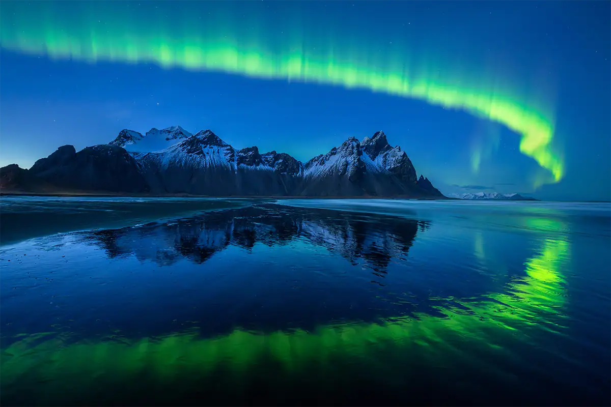 A aurora boreal brilhando sobre montanhas cobertas de neve e refletindo nas águas escuras da costa, fotografada com a lente zoom ultragrande angular Tamron 12-20 mm F2.8.