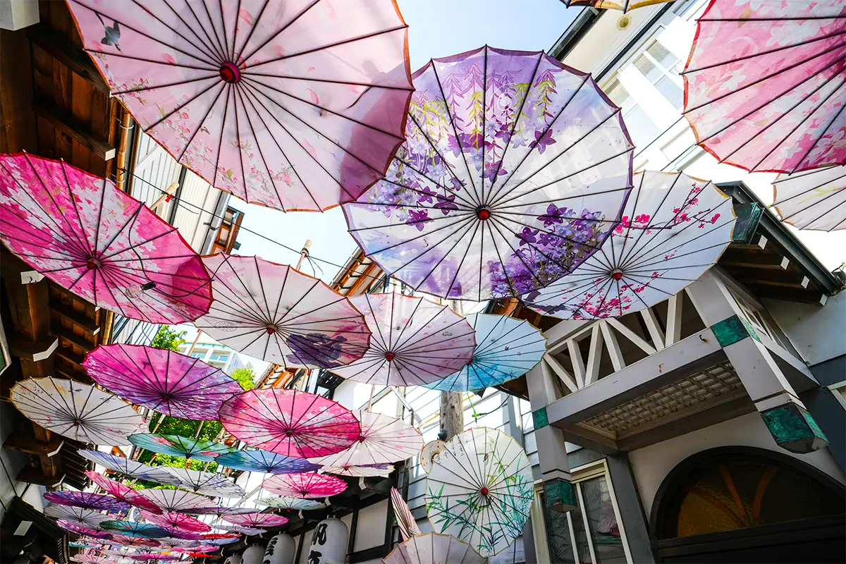 Fotografia de viagem em grande angular, tirada olhando para cima em uma rua estreita decorada com guarda-chuvas japoneses coloridos suspensos entre os prédios ao redor.
