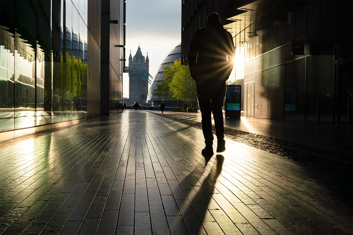 Fotografia de rua em grande angular de um pedestre em silhueta caminhando entre prédios modernos, com a Tower Bridge ao longe e a luz do sol criando longos reflexos na calçada.
