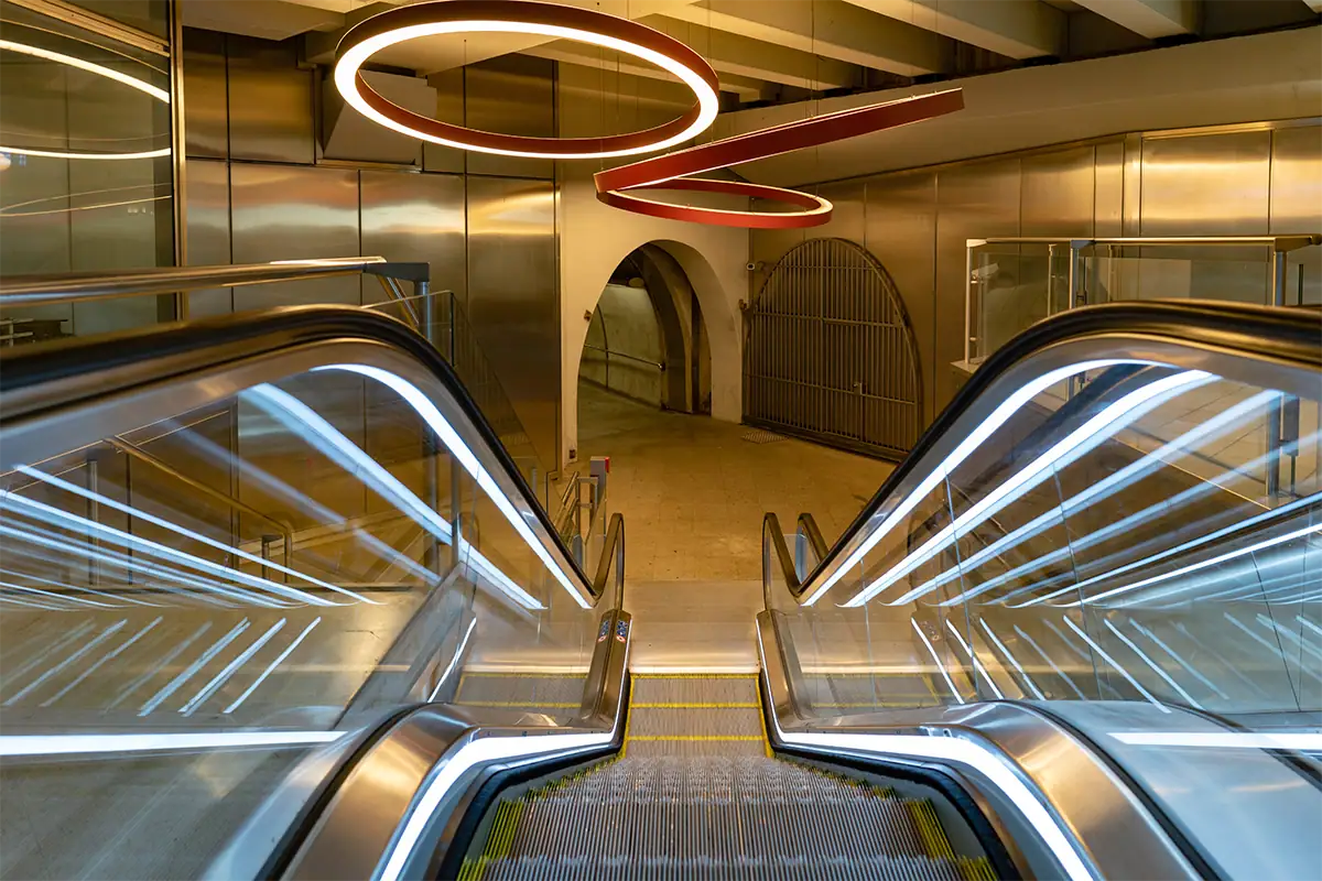 Fotografia de interior em grande angular de escadas rolantes modernas com corrimãos iluminados, luminárias circulares no teto e linhas arquitetônicas marcantes que conduzem o olhar pela cena.