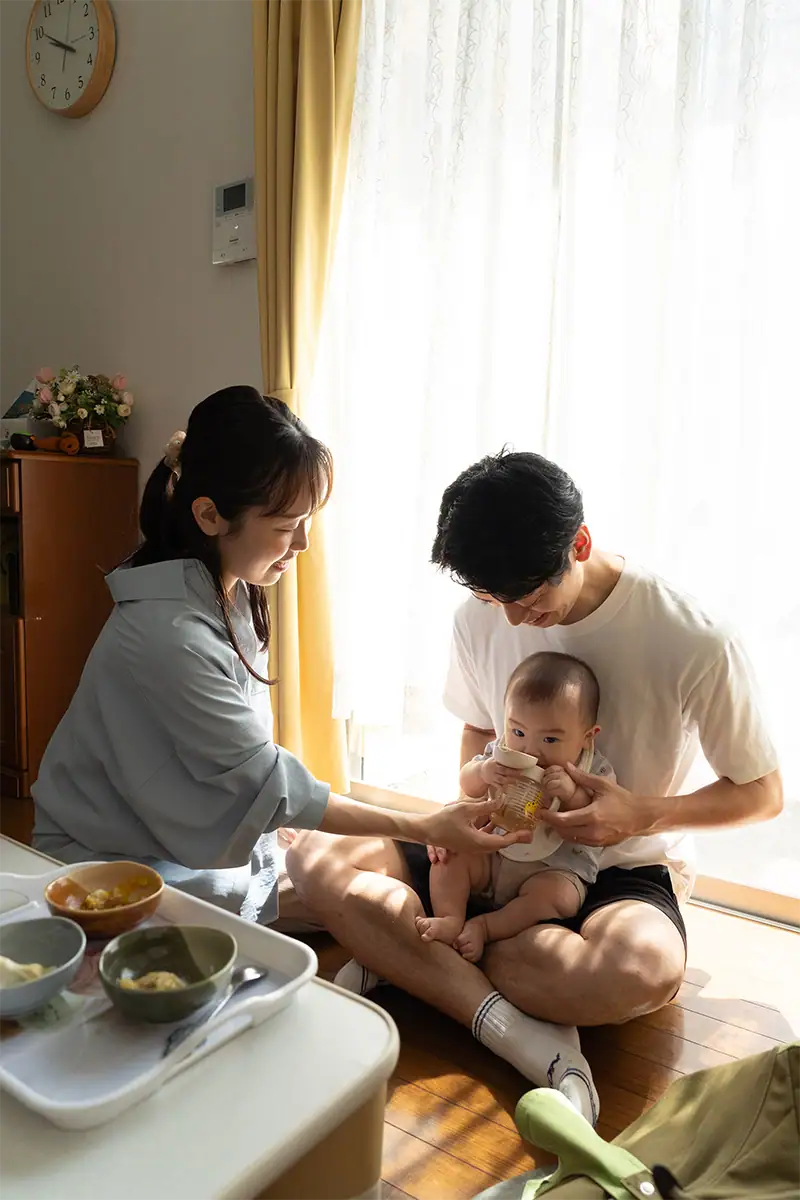 A mãe e o pai sentados com seu bebê ao lado de uma janela iluminada pelo sol, durante um momento cotidiano em família em casa.