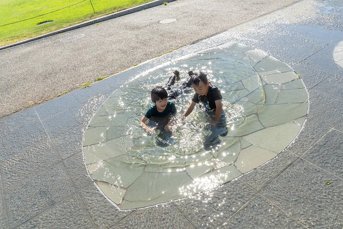 Duas crianças brincando em um parque aquático com reflexos cintilantes na água, fotografadas com a Tamron 25-200 mm F2.8-5.6 G2.