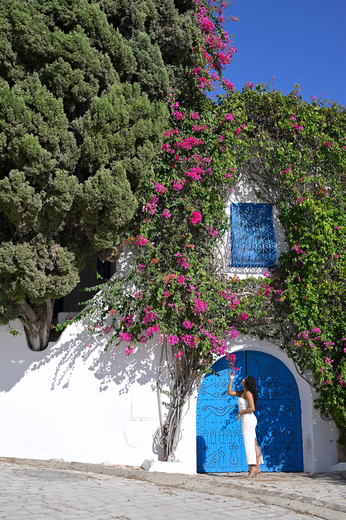 Woman in white standing beside a vivid blue door framed by pink bougainvillea and greenery on a sunlit white building.