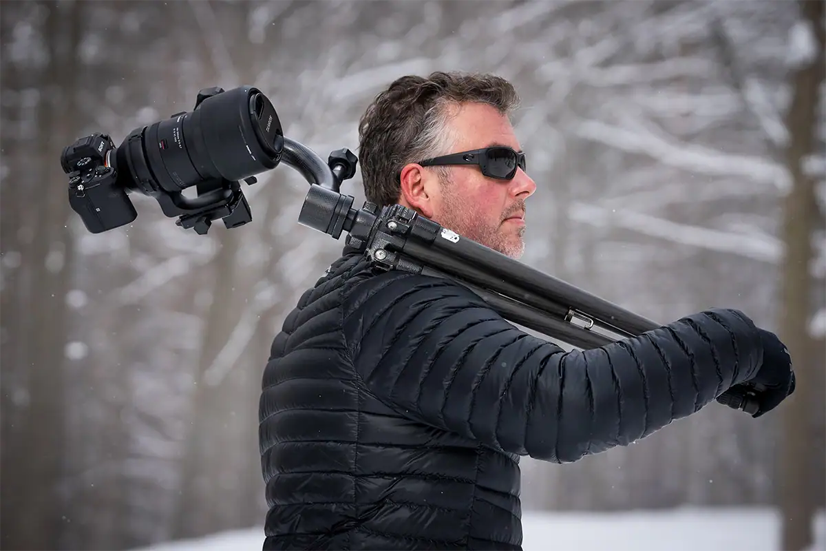 Photographer carrying a tripod with a gimbal head supporting a camera and Tamron telephoto lens in winter conditions.