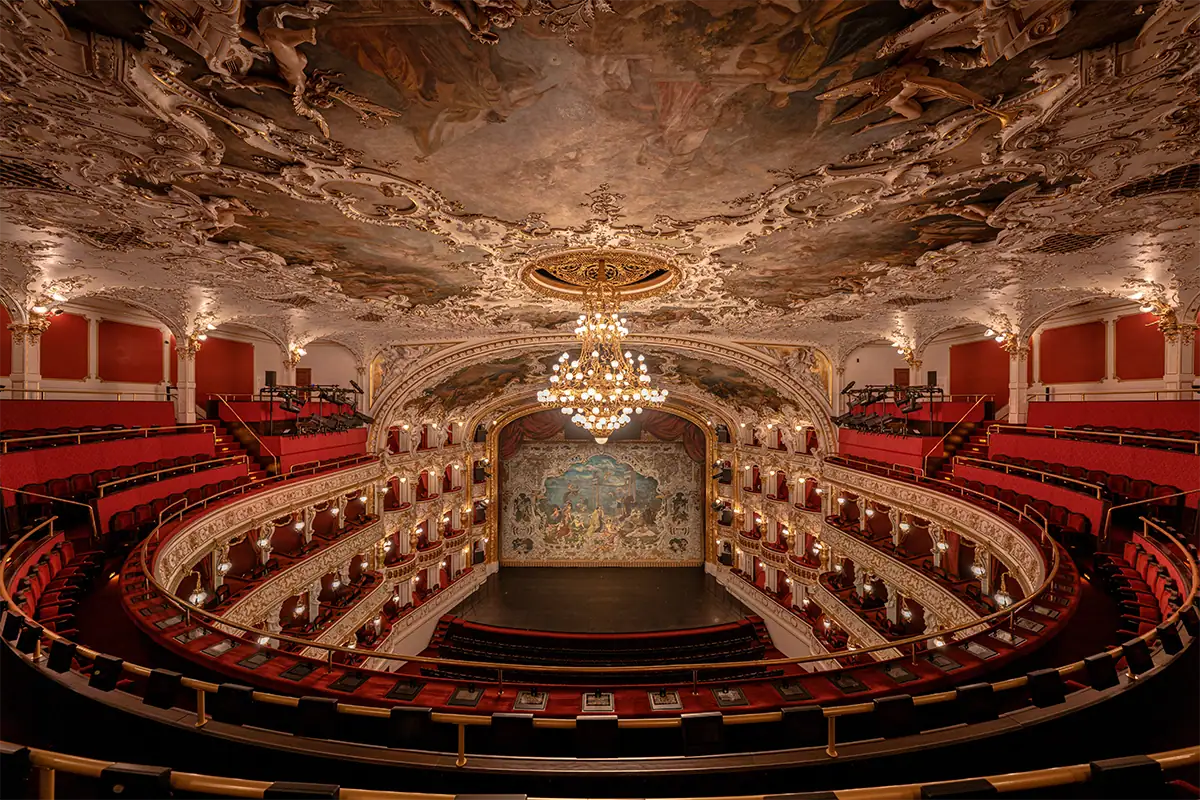 Fotografia de grande angular do interior de um teatro ornamentado, com assentos vermelhos, varandas curvas, lustres e pinturas detalhadas no teto.