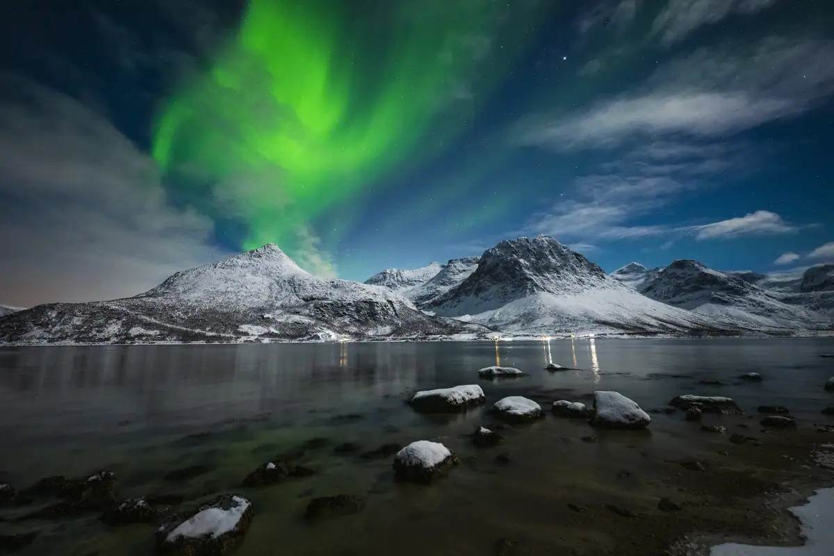 Paisagem noturna em grande angular mostrando a aurora boreal acima de montanhas cobertas de neve, águas calmas e rochas em primeiro plano.