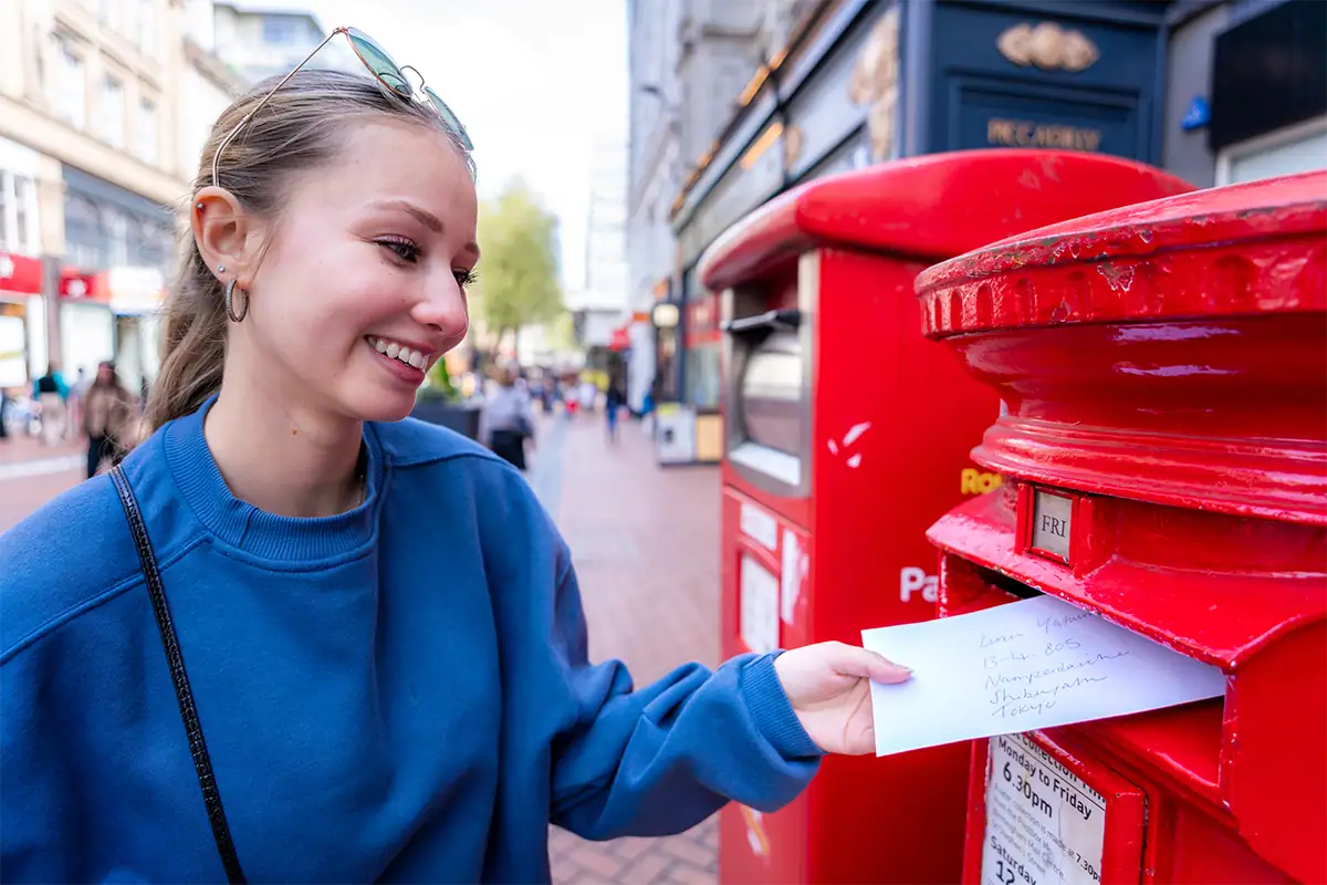 Retrato ambiental de uma mulher enviando uma carta ao lado de caixas de correio vermelhas brilhantes em uma rua da cidade, fotografado com uma lente grande-angular para incluir a personagem e o cenário urbano ao redor.