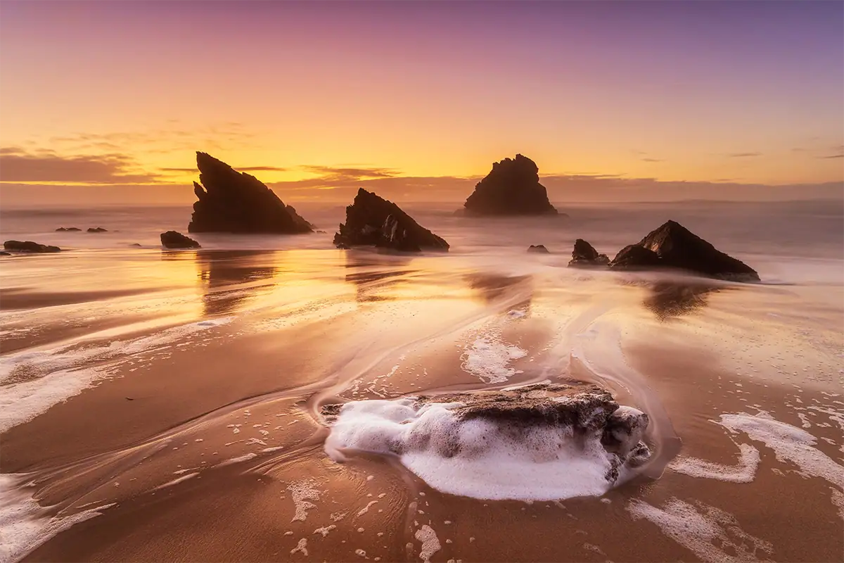 Paisagem costeira em grande angular ao pôr do sol, com formações rochosas no mar, areia molhada que reflete a luz e uma rocha em primeiro plano cercada pelas ondas que quebram.