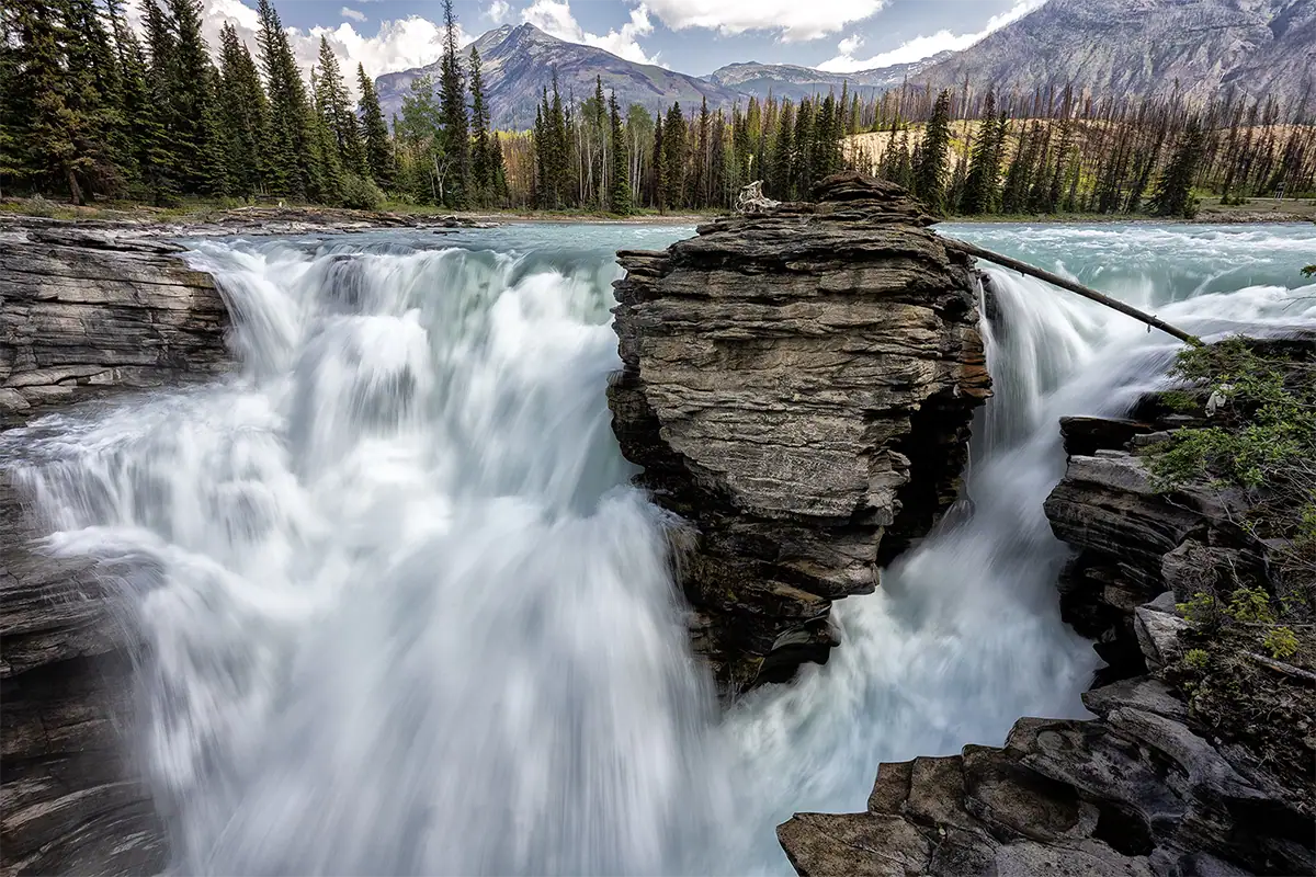 Paisagem em grande angular de uma imponente cachoeira de água turquesa que corre ao redor de uma grande rocha em primeiro plano, com uma floresta perene e montanhas ao longe.