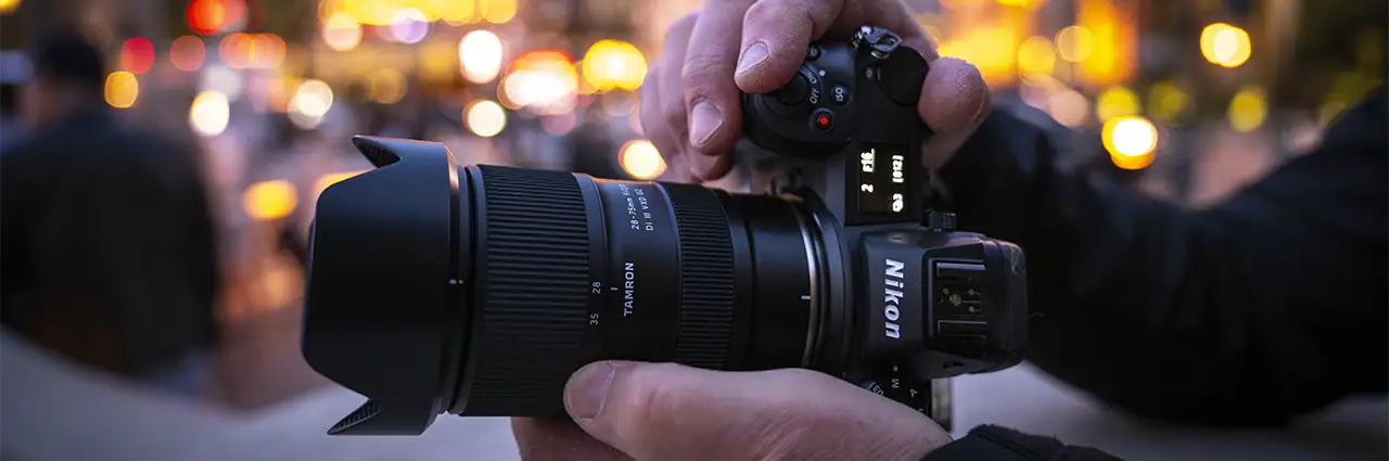 Close-up de um fotógrafo segurando uma câmera sem espelho da Nikon equipada com uma lente zoom grande-angular da Tamron, tendo como pano de fundo a cidade levemente desfocada ao entardecer.