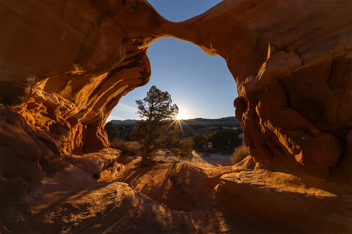 O nascer do sol brilhando através de um arco natural de arenito, em uma composição paisagística em grande angular, com o sol posicionado próximo à borda do quadro.