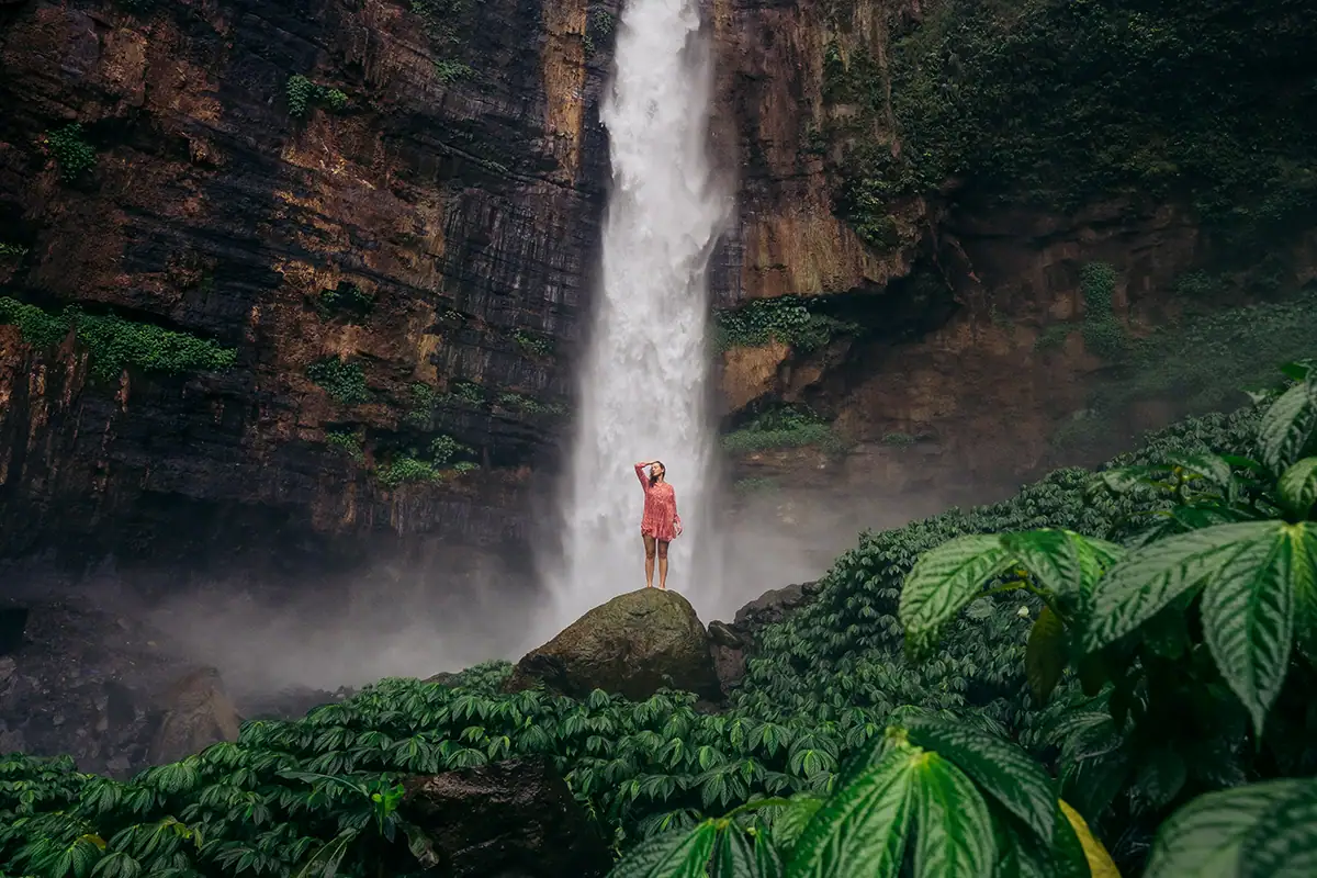 Uma mulher em pé diante de uma enorme cachoeira, com folhagem verde em primeiro plano