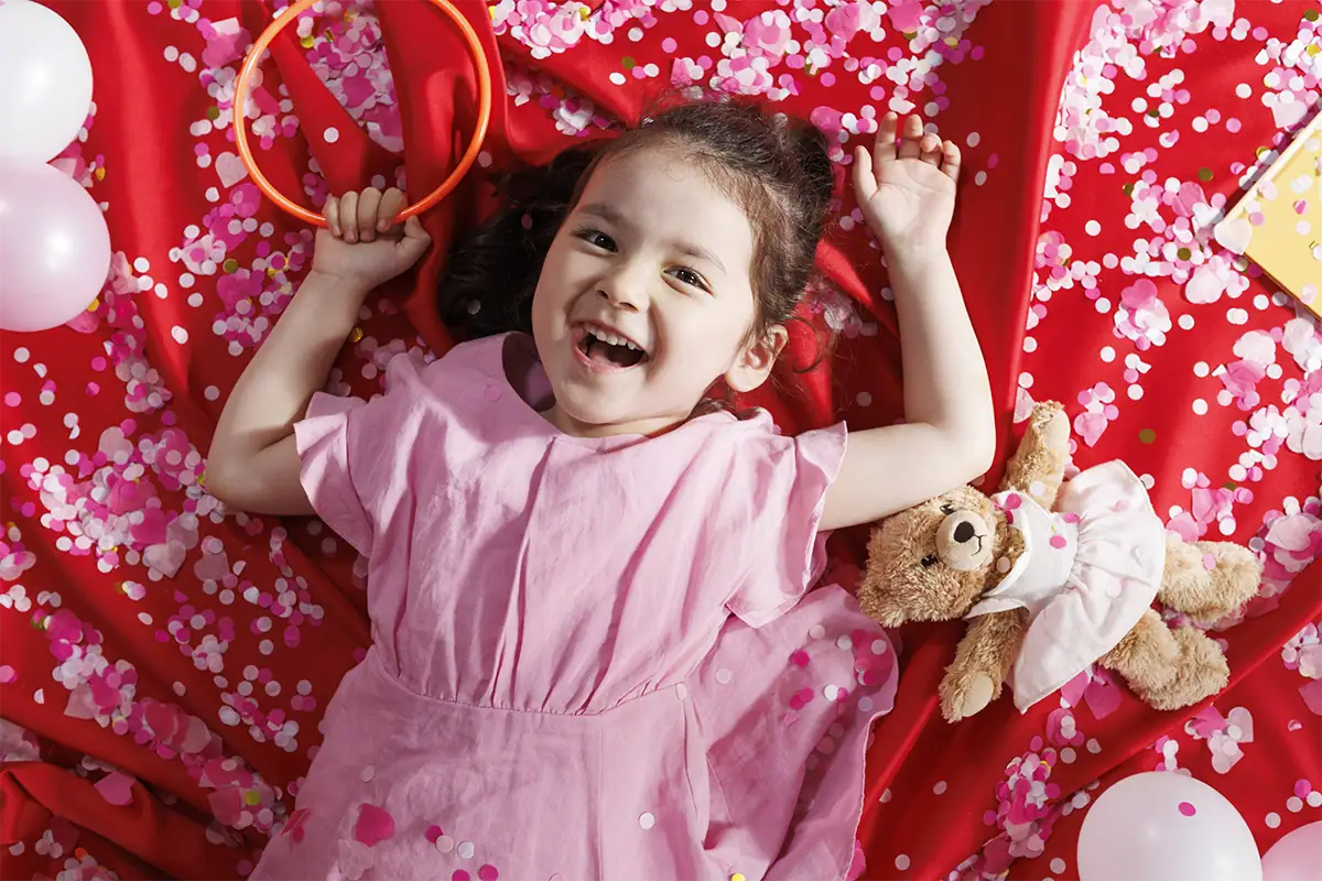 Laughing child photographed from above while lying among bright pink confetti, balloons, and birthday decorations.