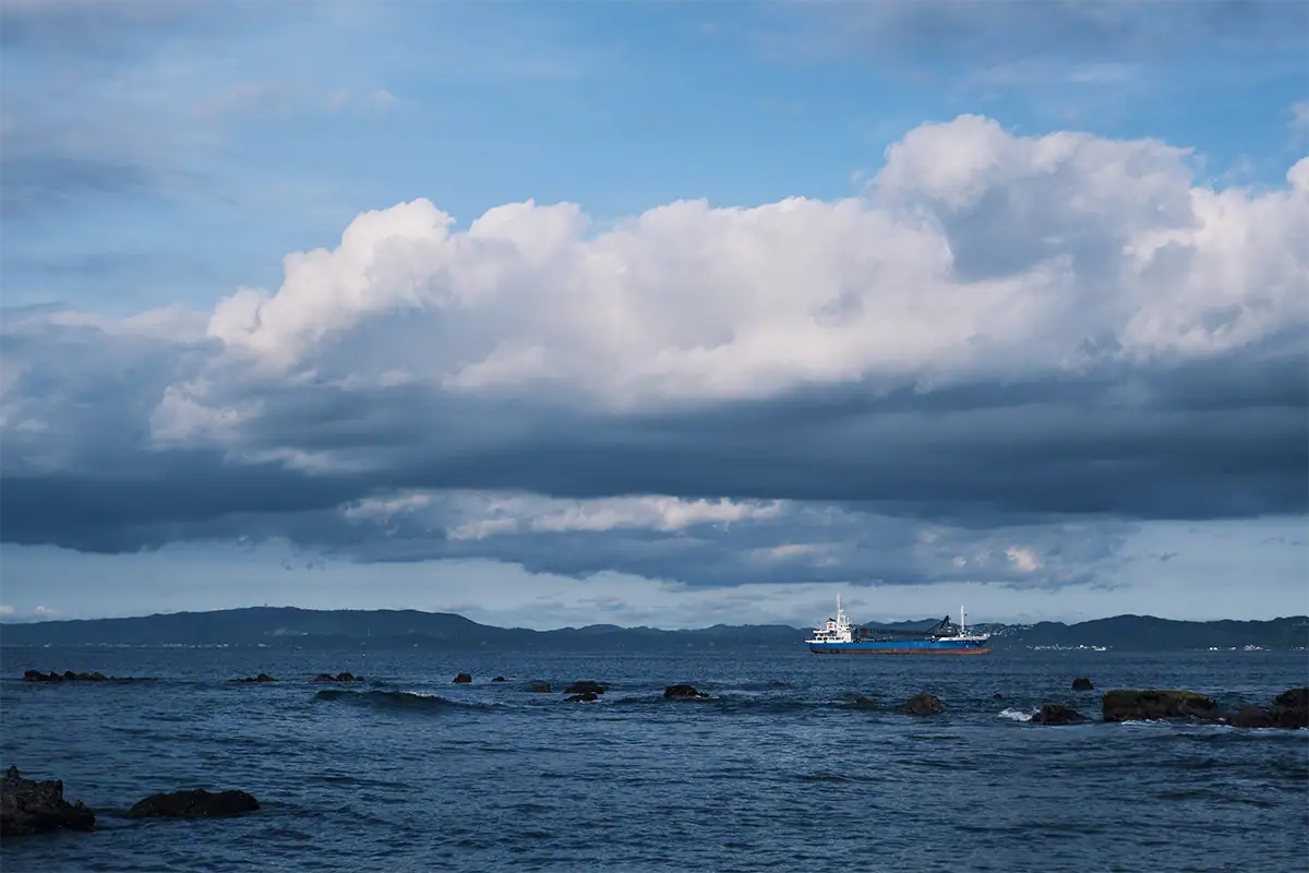 Dramatic layered clouds over dark blue coastal water with a cargo ship near the horizon.