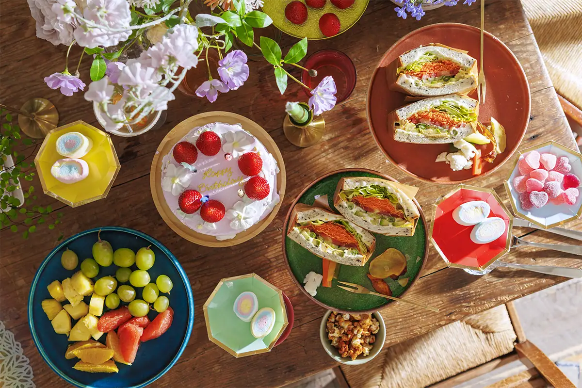 Overhead view of a colorful birthday table filled with cake, fruit, flowers, sandwiches, and other party foods.