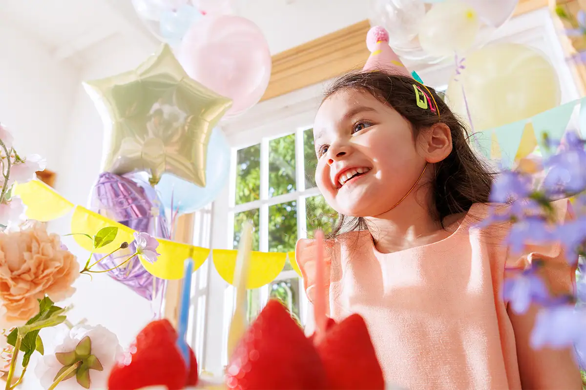 Smiling child seated at a colorful birthday table surrounded by balloons, flowers, and party decorations.