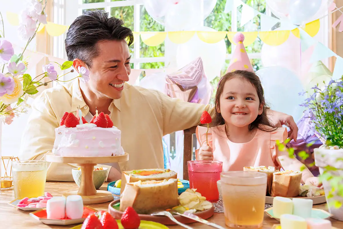 Father and daughter smiling together at a colorful birthday table with cake, flowers, drinks, and party decorations.