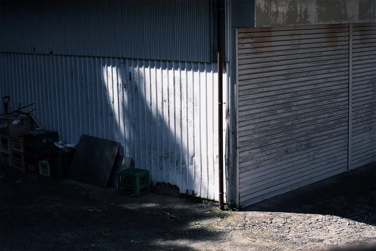 Sunlight falling across a white corrugated metal wall beside a dark garage door and stacked crates.