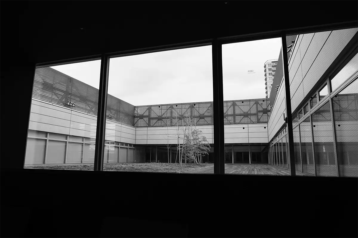 Black-and-white view through dark window frames toward a minimalist modern courtyard and surrounding architecture.