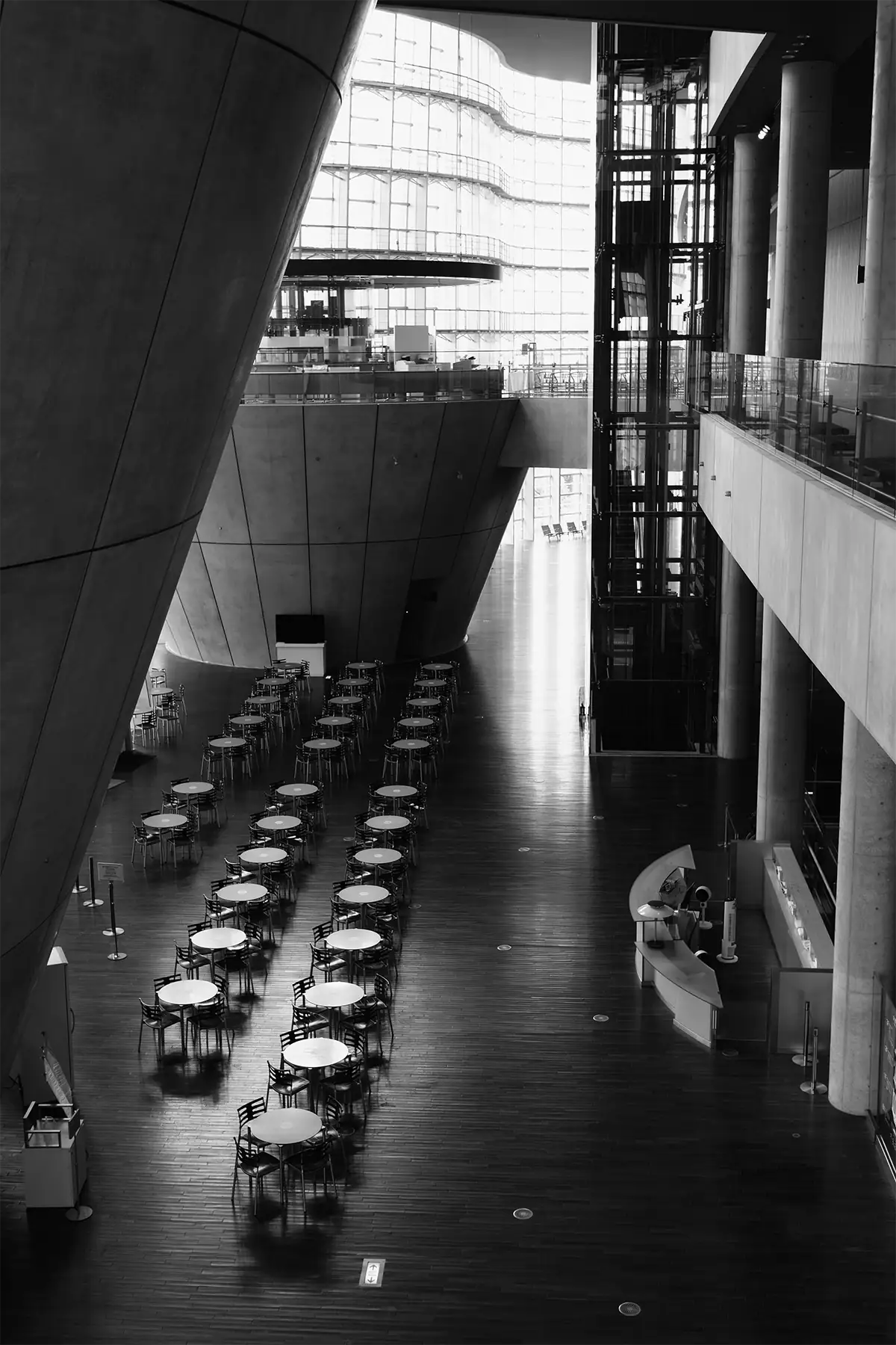 Black-and-white interior of The National Art Center, Tokyo, with rows of tables, curved concrete forms, glass walls, and elevators.