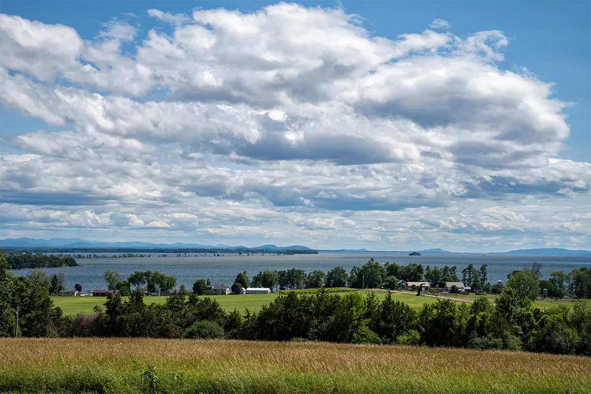View from a grassy hill in South Hero, Vermont, overlooking farms, Lake Champlain, distant mountains, and dramatic clouds.