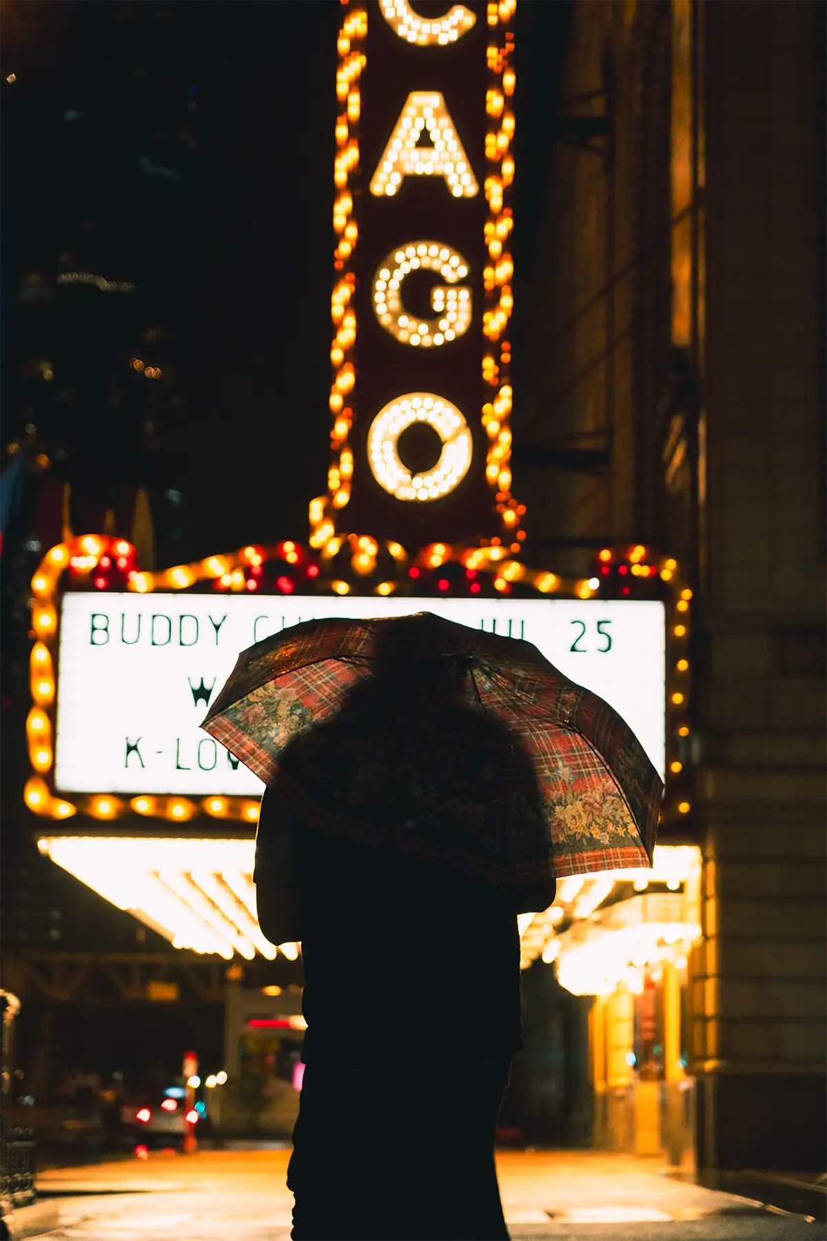 Pedestrian with an umbrella silhouetted beneath the illuminated Chicago Theatre marquee on a rainy night, photographed at 50mm.