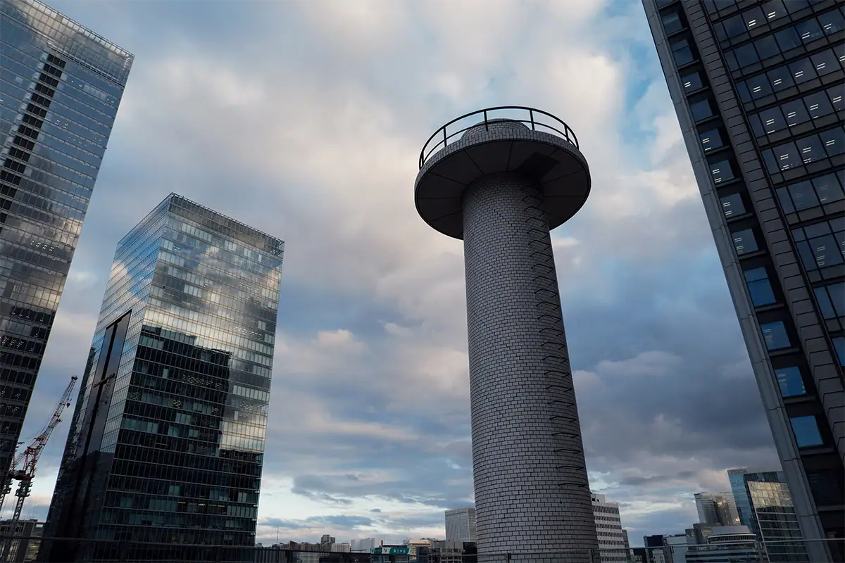Modern observation tower framed by glass office buildings beneath a dramatic cloudy sky.