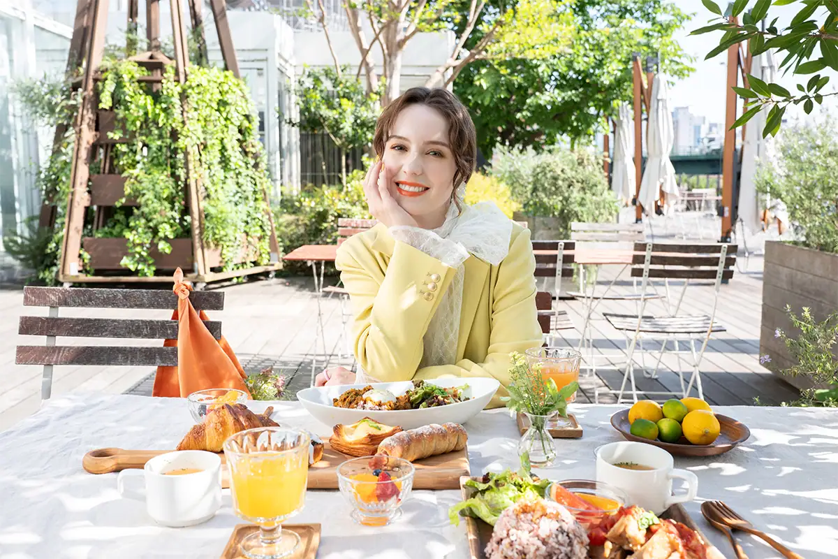 Mulher sorrindo em uma mesa de café ao ar livre, cercada por comida e vegetação, fotografada com uma distância focal de 34 mm usando a lente Tamron 20-40 mm F2.8.