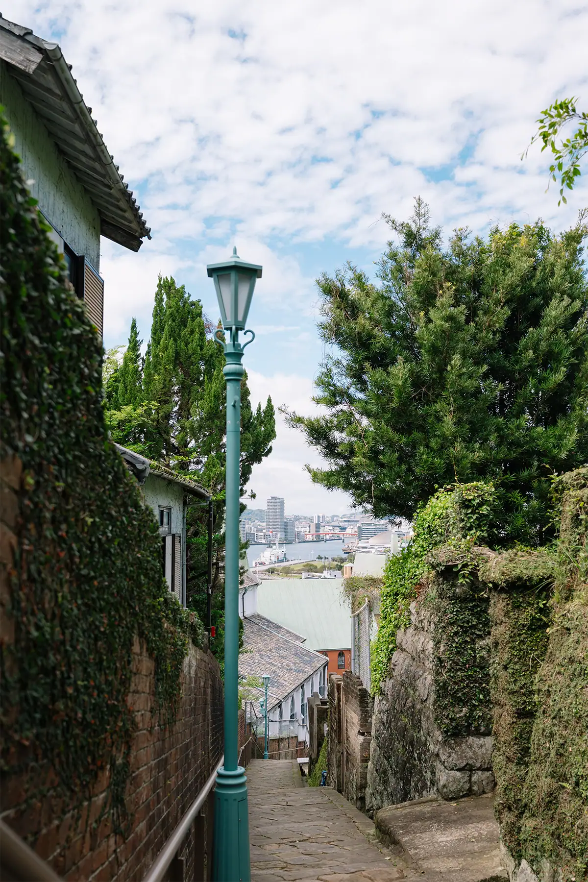 Stone alley descending toward a harbor with a green streetlamp and leafy walls, photographed at 36mm.