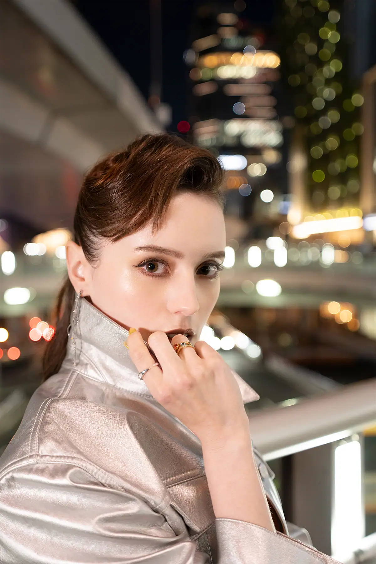 Retrato noturno de uma mulher vestindo uma jaqueta metálica, com as luzes coloridas da cidade e um bokeh circular ao fundo.