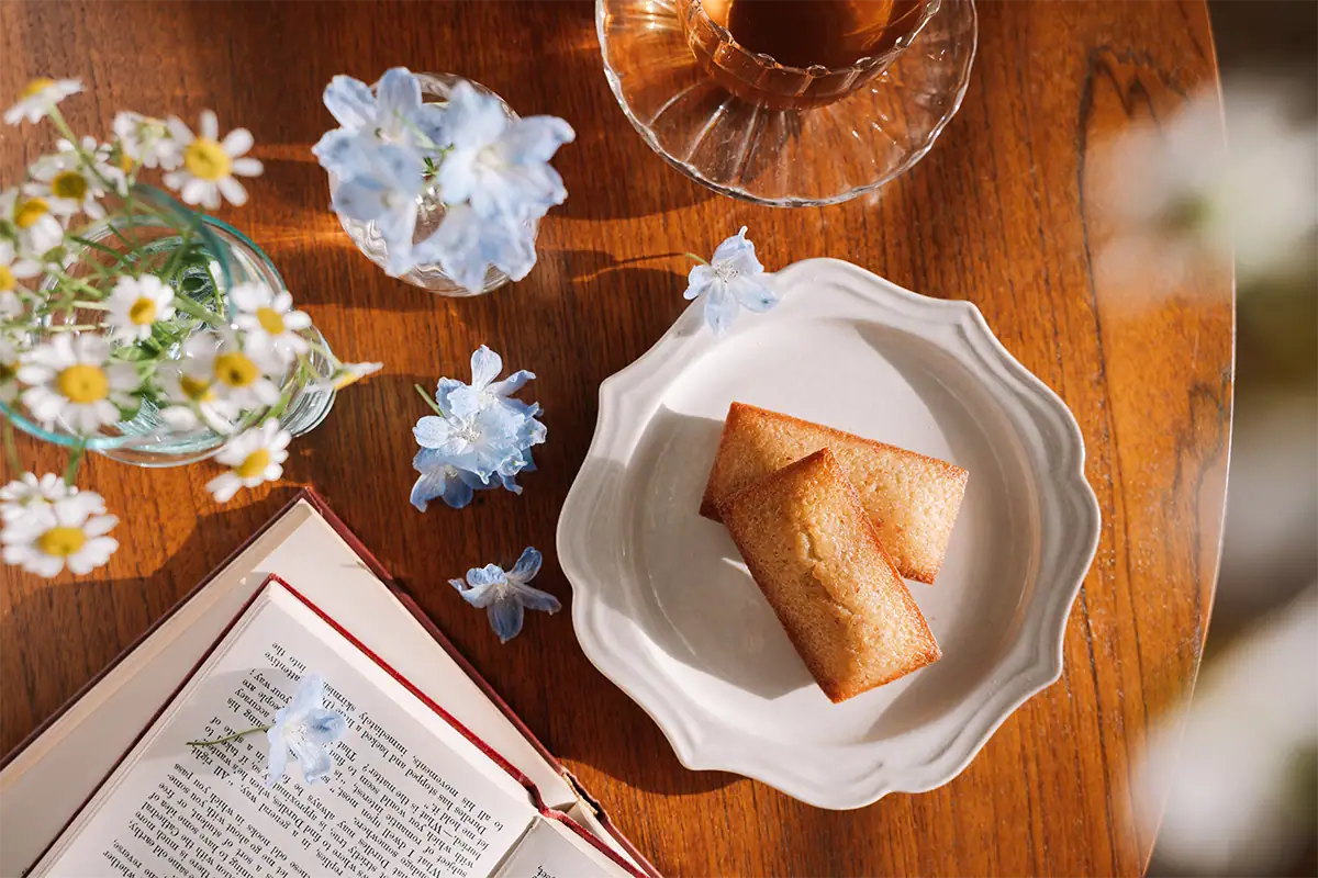 Overhead tabletop scene with baked goods, flowers, an open book, and tea in warm window light, photographed at 24mm.