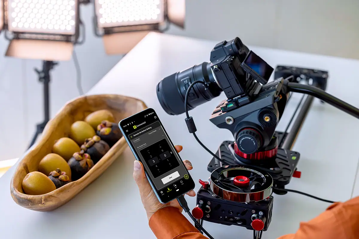 Smartphone équipé de l'application « TAMRON Lens Utility Mobile » à côté d'un appareil photo hybride muni d'un objectif Tamron, permettant de contrôler la mise au point en mode vidéo.