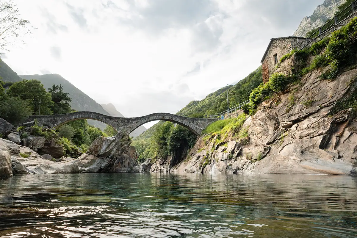 Un pont historique en pierre à double arche enjambe une rivière de montagne aux eaux turquoise, entourée de falaises rocheuses et de collines verdoyantes. Photo prise avec l'objectif Tamron 16-50 mm F2.8 A pour Nikon Z.
