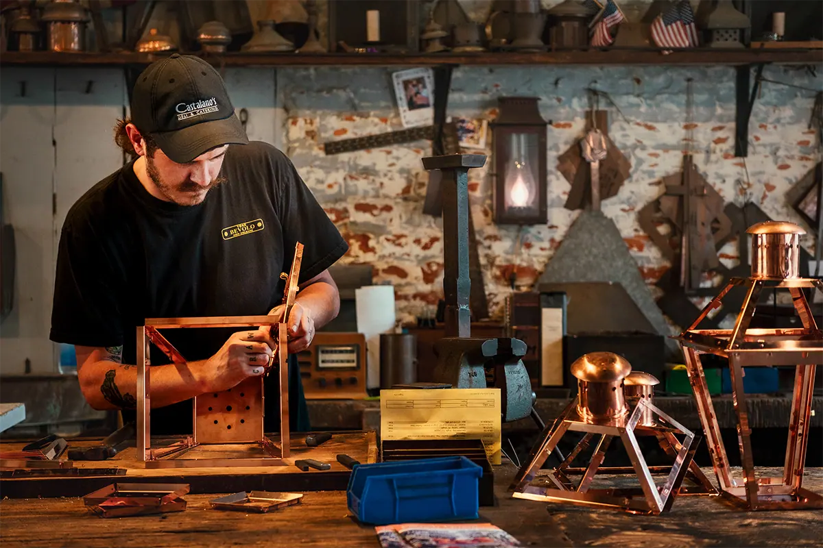 Un artesano con gorra y camisa negras trabajando en una linterna de cobre en un taller, fotografiado con el lente Tamron 16-50 mm F2.8 A para cámaras Sony con montura E y sensor APS-C.