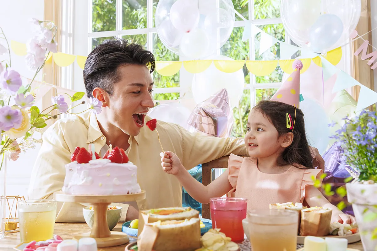 Un père et sa petite fille partagent un moment de joie autour d'un gâteau d'anniversaire à la fraise lors d'une fête haute en couleur organisée à l'intérieur, photographiée avec le Tamron 16-50 mm F2,8 A (B091).