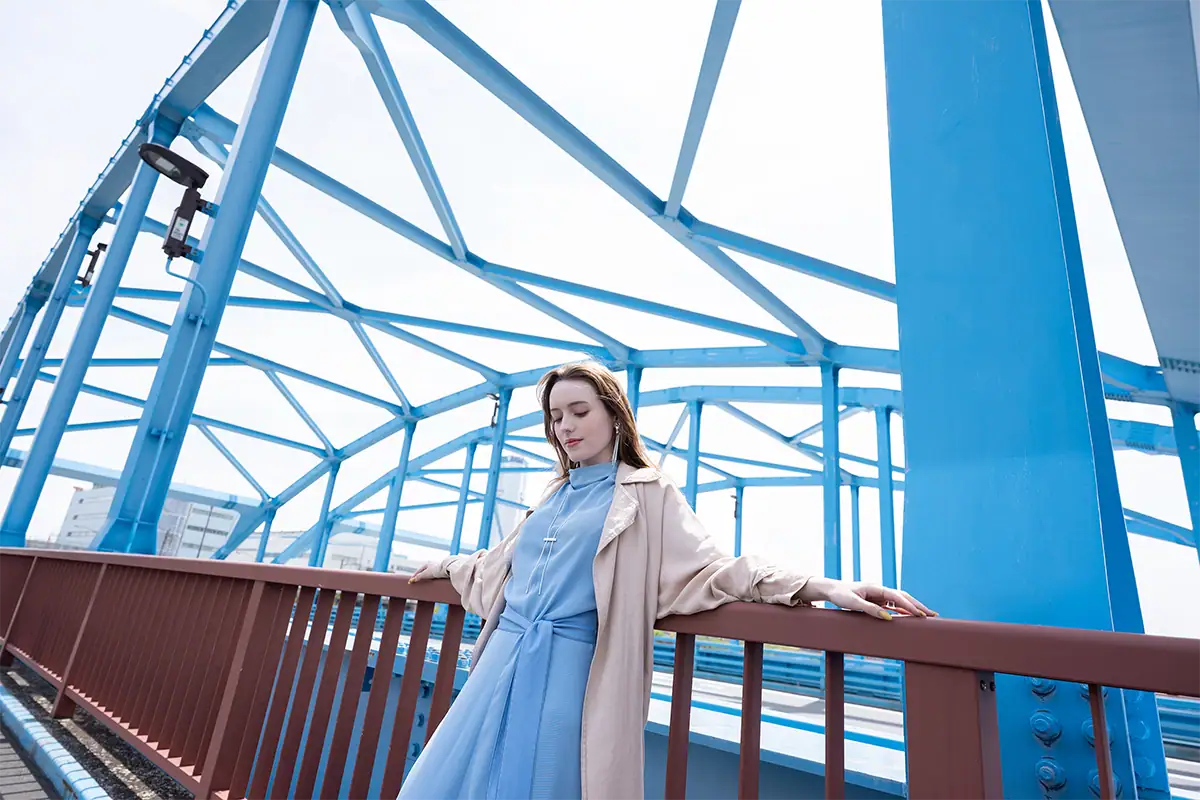 Femme en robe bleue posant sur un pont aux poutres en acier d'un bleu éclatant, photographiée avec l'objectif Tamron 20-40 mm F2,8 pour monture Nikon Z.