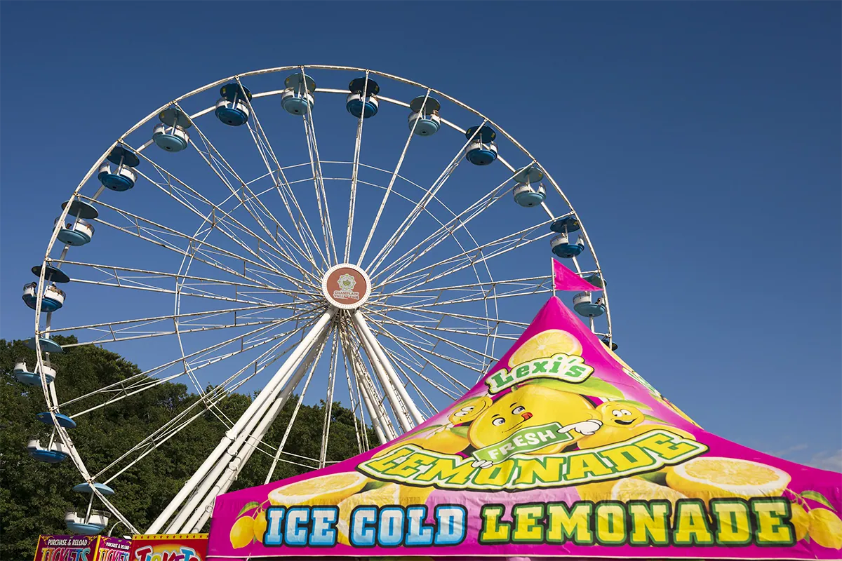 Une grande roue se dressant derrière un stand de limonade rose aux couleurs vives lors d'une fête foraine en plein air, sous un ciel bleu limpide, photographiée avec l'objectif Tamron 20-40 mm F2.8 pour monture Nikon Z.